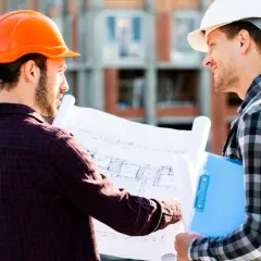 Dwóch inżynierów w kaskach omawia plany budowy. Ich praca obejmuje różne formy dozoru technicznego, zapewniając bezpieczeństwo i zgodność z projektem.