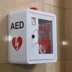 Biała obudowa AED z czerwonym sercem i symbolem błyskawicy, zamontowana na ścianie.