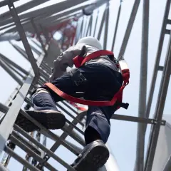 Pracownik z uprawnieniami wysokościowymi wspina się po drabinie masztu telekomunikacyjnego, zabezpieczony uprzężą.