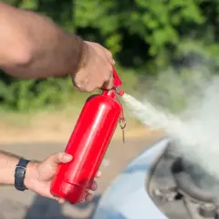 Ręce gaszą pożar pod maską samochodu czerwoną gaśnicą. To ważny element wyposażenia, bo nigdy nie wiadomo, jaka gaśnica do samochodu będzie potrzebna w awaryjnej sytuacji.