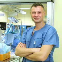 Lekarz w niebieskiej koszuli chirurgicznej stoi na tle sali operacyjnej.