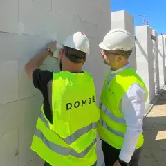 Dwóch mężczyzn w kaskach i kamizelkach odblaskowych omawia postęp prac na budowie. Dbają o bhp, sprawdzając detale konstrukcji z bloczków.