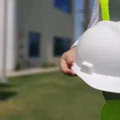Biały kask budowlany w dłoni, obok jaskrawożółtej kamizelki. Różne kolory kasków na budowie to symbol bezpieczeństwa.