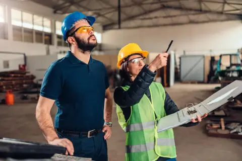Inspektor BHP w żółtym kasku i kamizelce omawia z kolegą plany na hali produkcyjnej.