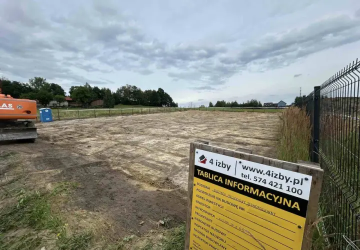 Plac budowy z koparką i toaletą przenośną. Na pierwszym planie tablica informacyjna z napisem "TABLICA INFORMACYJNA".