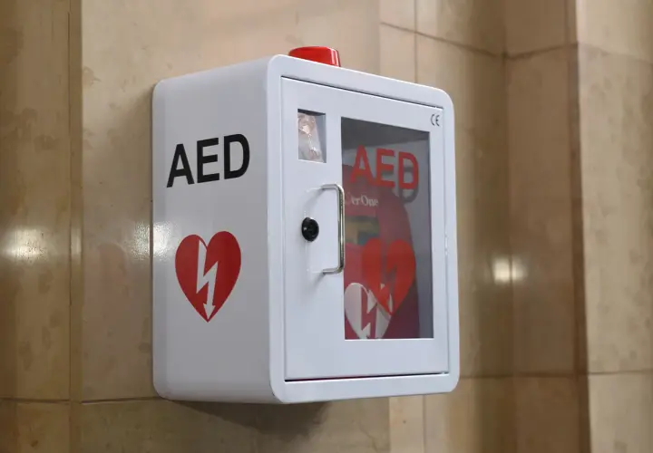Biała obudowa AED z czerwonym sercem i symbolem błyskawicy, zamontowana na ścianie.