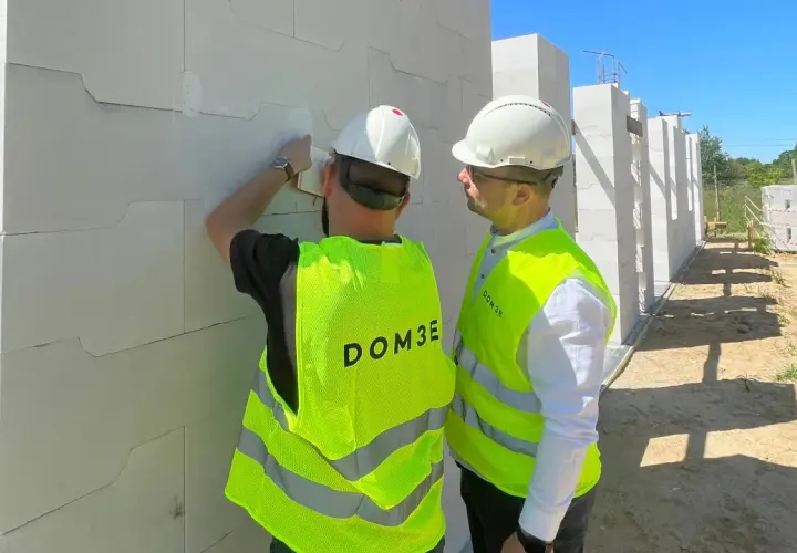 Dwóch mężczyzn w kaskach i kamizelkach odblaskowych omawia postęp prac na budowie. Dbają o bhp, sprawdzając detale konstrukcji z bloczków.