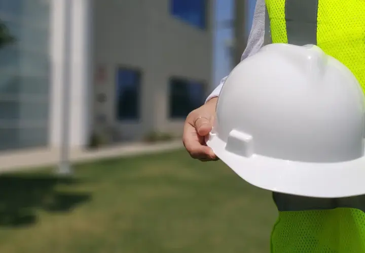 Biały kask budowlany w dłoni, obok jaskrawożółtej kamizelki. Różne kolory kasków na budowie to symbol bezpieczeństwa.