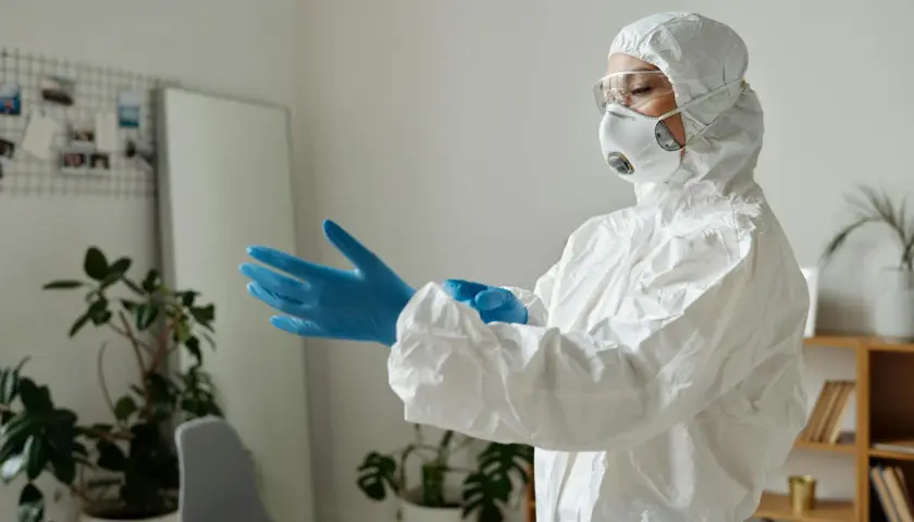 Osoba w kombinezonie ochronnym zakłada niebieskie rękawiczki, przygotowując się do pracy z czynnikami biologicznymi.
