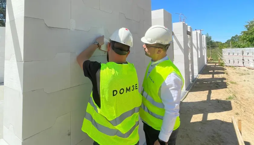 Dwóch mężczyzn w kaskach i kamizelkach odblaskowych omawia postęp prac na budowie. Dbają o bhp, sprawdzając detale konstrukcji z bloczków.