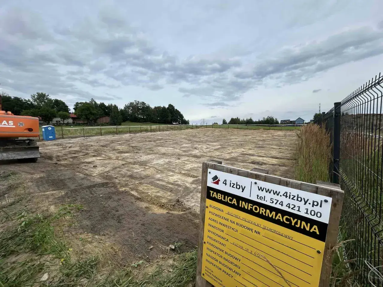 Plac budowy z koparką i toaletą przenośną. Na pierwszym planie tablica informacyjna z napisem "TABLICA INFORMACYJNA".