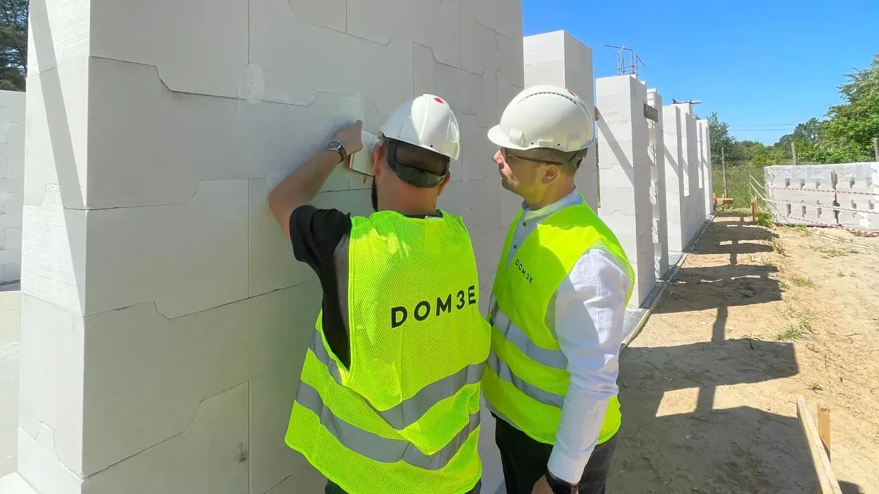 Dwóch mężczyzn w kaskach i kamizelkach odblaskowych omawia postęp prac na budowie. Dbają o bhp, sprawdzając detale konstrukcji z bloczków.