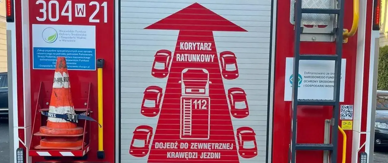 Na drzwiach wozu strażackiego widnieje grafika "KORYTARZ RATUNKOWY" z samochodami i numerem 112.