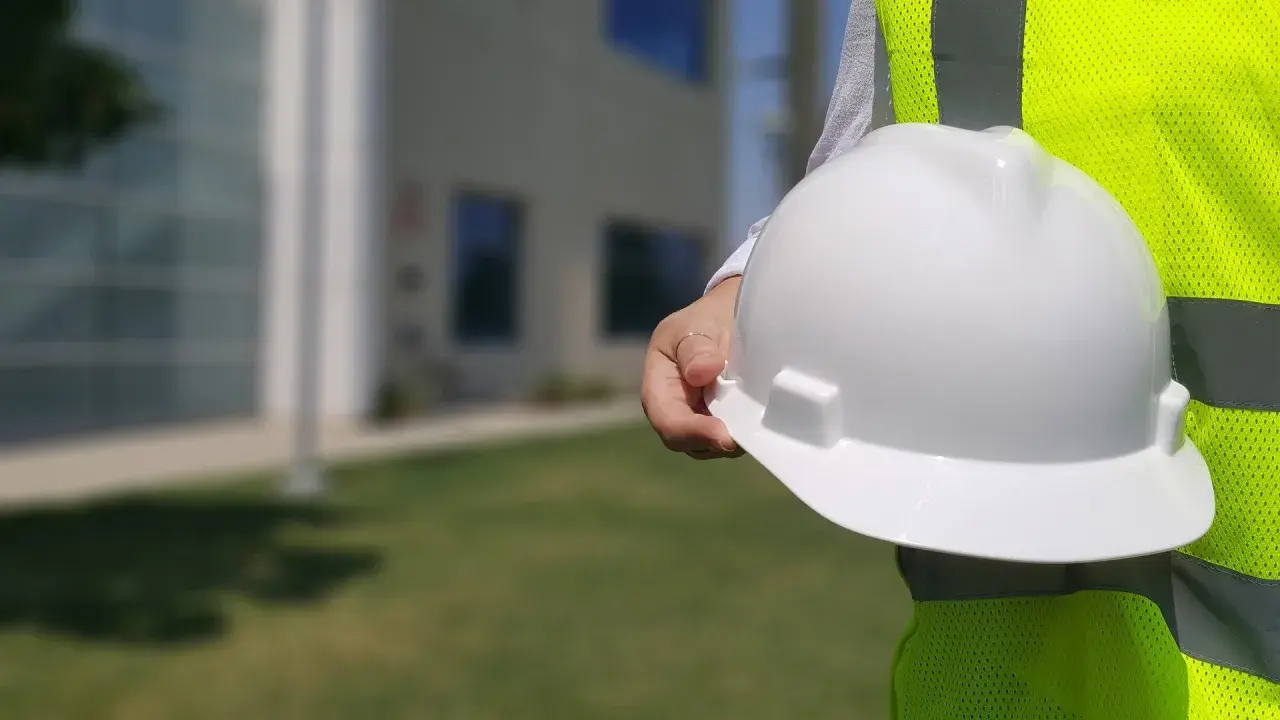 Biały kask budowlany w dłoni, obok jaskrawożółtej kamizelki. Różne kolory kasków na budowie to symbol bezpieczeństwa.